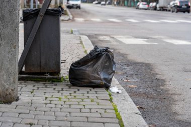 Sabahın erken saatlerinde bir şehir caddesinde geri dönüşüm kutusunun yanındaki kaldırımda bir yığın atılmış siyah çöp torbası duruyor..