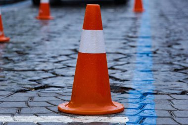 Yağmura batırılmış kaldırım taşı caddesinde, kentsel alanda yaya güvenliği için inşaat alanı oluşturan parlak turuncu trafik konileri yer alıyor..