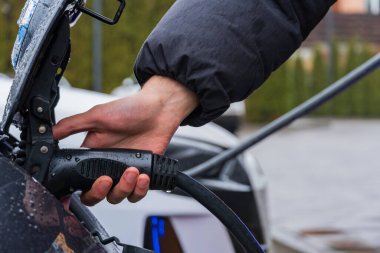 Bir el elektrik kablosunu elektrikli bir araca bağlıyor, teknolojinin ve sürdürülebilir ulaşımın karışımını şehir ortamında gösteriyor.