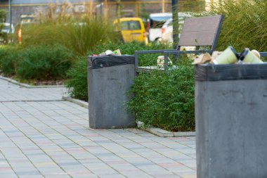 Çöp bidonları bir park bankının yanında, yeşilliklerle çevrili ve kalabalık bir şehir ortamında kaldırım taşlarıyla çevrili..