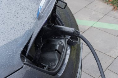 Şarj kablosu elektrikli aracın limanına bağlanırken, şehir merkezinde arabanın yüzeyinde yağmur damlaları birikiyor..