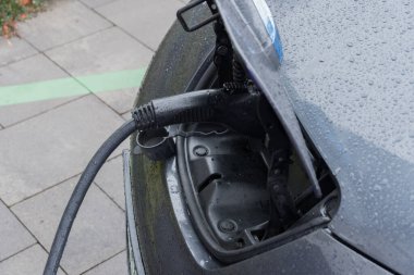 Elektrikli bir araç, halka açık bir şarj istasyonuna bağlıdır. Arabanın üzerinde yağmur damlaları ve şehrin çevresindeki kaldırımlar vardır..