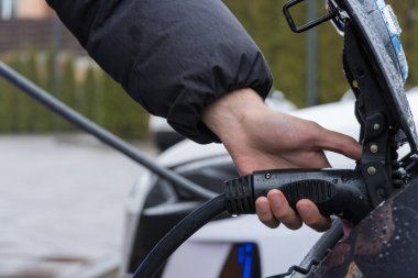Bir insan şarj kablosunu bir şarj istasyonundaki bir elektrikli araca bağlar. Çiseler ve bulutlu gökyüzü tepemizde belirir..
