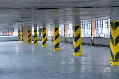 Geniş otopark sarı ve siyah çizgili kolonlar, geniş pencereler doğal ışık ve temiz beton zemin içerir..