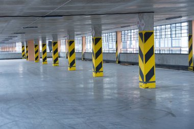 Boş bir park alanında modern bir tasarım sergilenen koyu sarı ve siyah çizgili destek kolonları bulunur. Doğal ışık büyük pencerelerden süzülür.