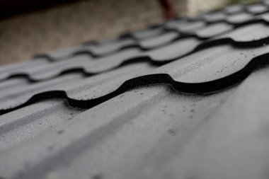 Su damlacıkları desenli bir metal çatı yüzeyinde dinleniyor. Kıvrımlı tasarımını sergiliyor ve yakın zamanda yağan yağmurun ardından parlak bir şekilde tamamlanıyor..