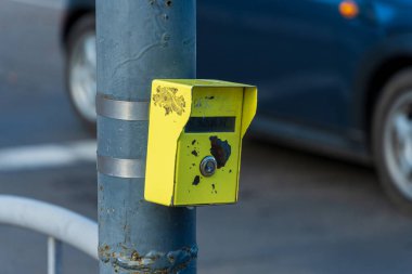 Yıpranmış bir direk parlak sarı bir trafik ışığı düğmesini destekliyor, kalabalık bir şehir caddesinde bir araba geçiyor..