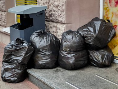 Yoğun bir şehir kaldırımındaki bir dükkanın önündeki çöp kutusunun yanına siyah çöp torbaları istiflenir..