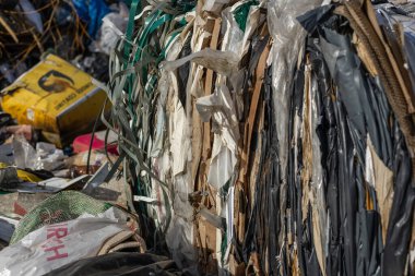 Yığılmış plastik ve karton atık balyaları, gün ortası güneş ışığıyla aydınlatılan bir geri dönüşüm merkezinde görülür..