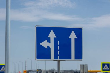 Bir yol işareti, mavi gökyüzünün altındaki bir kavşakta sürücüleri yönlendiren düz ve solu gösteren okları gösterir..