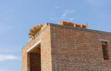 Tuğladan bir yapı, çatısına yerleştirilmiş ahşap kirişlerle tamamlanmamış, parlak mavi gökyüzünün altında, devam eden inşaat çalışmalarını gözler önüne seriyor..