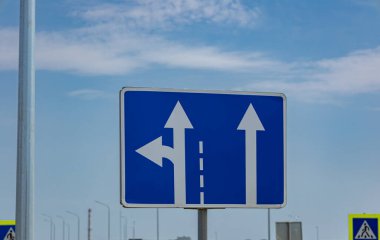 Trafik levhası, üzerinde mavi gökyüzü olan bir yolda araçlar için sol ve düz yönleri gösteren oklar gösteriyor..