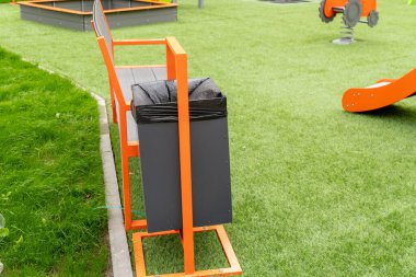 Siyah bir çöp tenekesinin yanındaki parlak turuncu bank güneşli bir günde canlı bir halk parkında temizliği ön plana çıkarıyor..