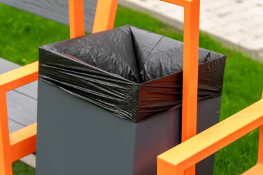 Siyah silindir ve parlak turuncu çerçeveli çöp tenekesi güneşli havalarda kentsel alan temizliğini vurgulayarak bir park bankının yanında oturur..