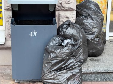 Kentsel bölgelerdeki atık yönetimi sorunlarını vurgulayarak, bir şehir ortamında taşan bir çöp kutusunun yanına siyah çöp torbaları yığılıyor.