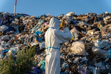 Beyaz tehlikeli madde giysisi içindeki bir kişi, parlak sabah saatlerinde çöp sahasında büyük bir atık yığınının yanında örnekler topluyor..