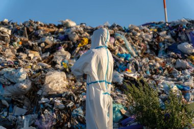 Bir çevre çalışanı, açık mavi gökyüzünün altında koruyucu giysiler içinde, kirliliğe odaklanmış, yüksek bir atık yığınını değerlendiriyor..