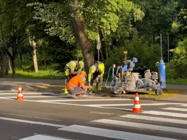 Şehir parkındaki ağaçlarla çevrili sakin bir yolda dört işçi yaya geçidine taze boya sürüyor. Takım çalışması ve çabalarını gösteriyor..