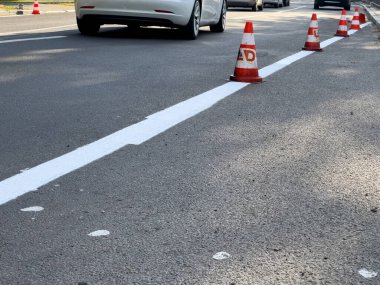 Trafik konileri, asfalt üzerinde yeni boyanmış beyaz bir çizgiyi işaret ediyor. Bu da, gün boyunca devam eden bir şehir ortamında yol çalışması olduğunu gösteriyor..