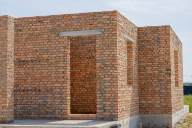 Açık bir alanda tuğla bina inşaatı devam ediyor. Duvarlar açık ve güneşli bir gün..