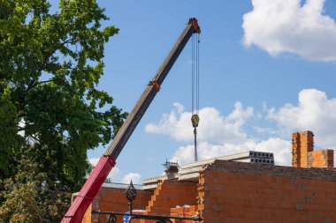 Büyük bir vinç inşaat sahasında çalışır, arka planda mavi gökyüzüne ve yeşil ağaçlara karşı inşaat malzemeleri taşır..