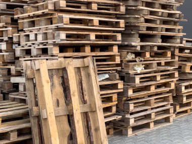 Büyük bir kullanılmış ahşap palet yığını birbiri üzerine yığılmış halde gündüz saatlerinde bir deponun yanında duruyor..