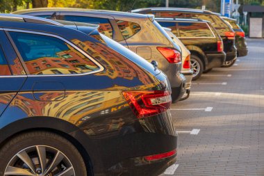 Şehir parkına çeşitli arabalar park edilmiş. Parlak güneş ışığı altında yakındaki binaların renkli yansımalarını sergiliyorlar..