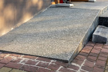 Geniş beton bir rampa, öğleden sonra güneşiyle aydınlanan tuğla bir yola bir bina girişini bağlar..