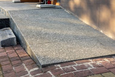 Gri taştan bir tekerlekli sandalye rampası, yapıyı tamamlayan tuğla geçidin yanındaki kamu binasına çıkıyor..