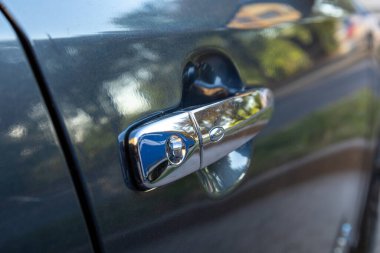 Parlak bir arabanın kapı kolunun yakın çekimi güneş ışığını yansıtıyor. Şık tasarımı ve banliyö evinin canlı çevresini gösteriyor..