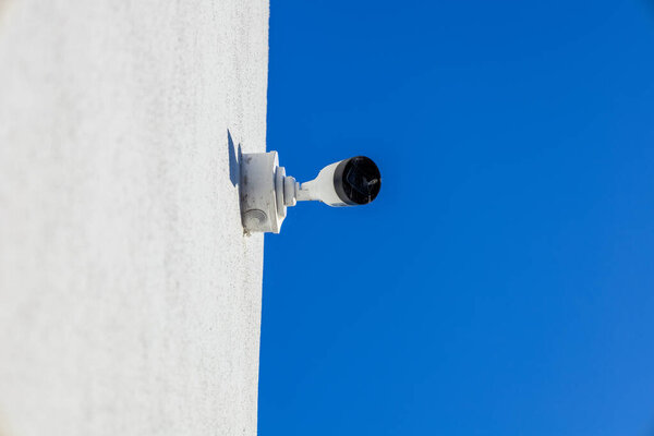A security camera is installed on a white structure, angled toward a vibrant blue sky, ensuring a watchful eye on its surroundings.
