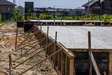 Workers are preparing a concrete foundation at a construction site surrounded by residential buildings under clear blue skies.