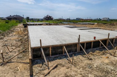 Workers are pouring concrete for a foundation at a construction site on a clear day, surrounded by new homes and green fields.