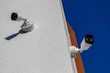 İki dış güvenlik kamerası beyaz bir duvara monte edilmiş, açık mavi gökyüzünün altındaki bölgeyi gün boyunca izliyorlar..