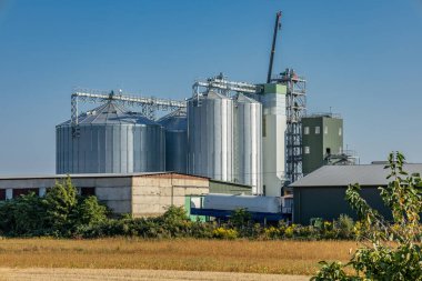 Grain silos stand tall in a rural landscape, while a crane works on construction in clear skies and vibrant surroundings.