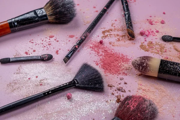Fashionable cosmetic background with decorative scattered and crumbled cosmetics,powder,blush,eyeshadows with sparkles and makeup brushes on a pink background.