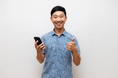Asyalı pozitif adam mavi gömlek elinde akıllı telefon gülümsemesi ve izole edilmiş baş parmağını göster.