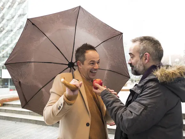 Two forty year old executives with umbrellas offer each other a red apple on a rainy winter street symbolizing mutual respect and camaraderie