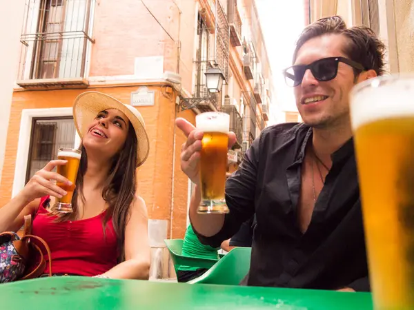 Latin kökenli, beyaz, güneş gözlüklü, diş telli, şapkalı, siyah tişörtlü, bira içerek gülen bir çift.