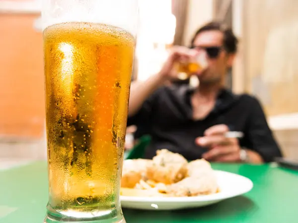 glass of beer on Spanish bar terrace with defocused caucasian man drinking and with cigar and sunglasses in background and fried fish appetizer