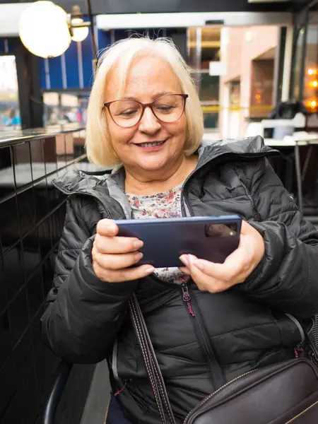 stock image blonde gamer grandmother with glasses very happy playing on smartphone in winter on a restaurant terrace