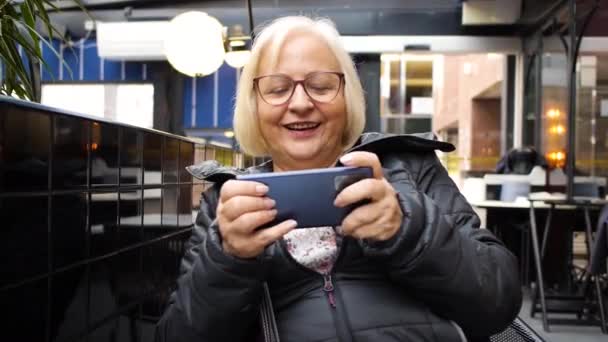 今年冬天 快乐的金发游戏玩家奶奶在餐厅的平台上玩智能手机 没有相机的动作 — 图库视频影像