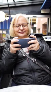 Kışın bir kafede akıllı telefondan video oyunu oynayan dikey büyükanne oyuncu.