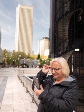 senior blonde business woman looks at cell phone holding glasses checking her vision on the street in the daytime in winter clipart