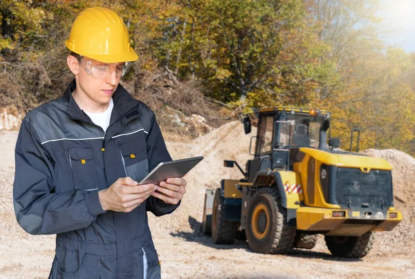 Engineer in a helmet with a digital tablet on the background of construction machine