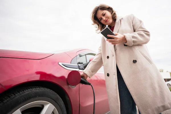 Akıllı Telefonlu Bir Kadın Kırmızı Elektrikli Arabayı Şarj Ediyor — Stok fotoğraf