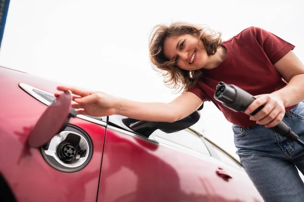 赤い電気自動車を充電スマートフォンを持つ女性 — ストック写真