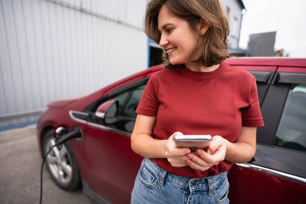 Akıllı Telefonlu Bir Kadın Kırmızı Elektrikli Arabayı Şarj Ediyor — Stok fotoğraf