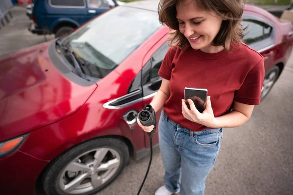 Жінка Смартфоном Заряджає Червоний Електромобіль — стокове фото