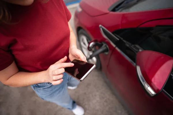 赤い電気自動車を充電スマートフォンを持つ女性 — ストック写真